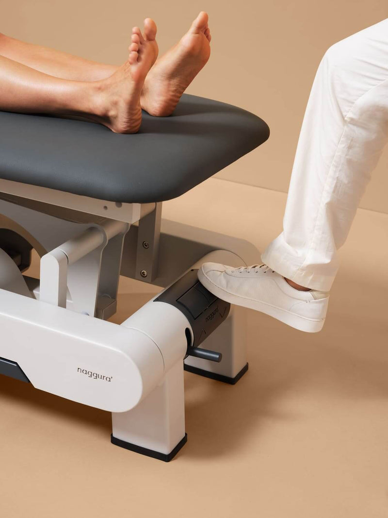 Therapeut betätigt Rundumschaltung an der Rückseite mit dem Fuß zur Höhenverstellung der Osteopathie-Liege Naggura N'RUN2W